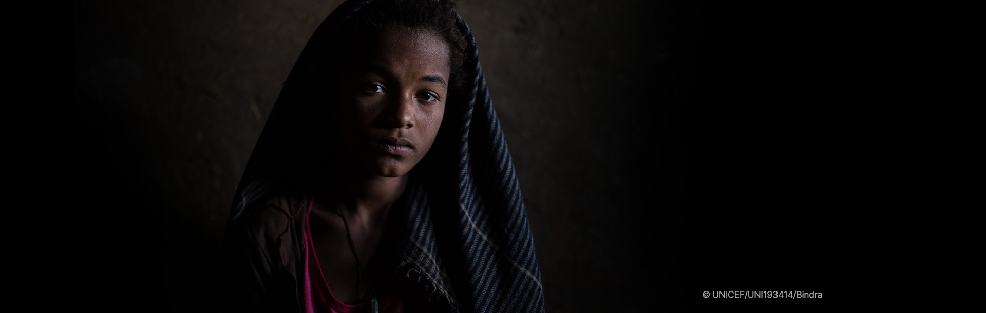 어두운 배경 속에서 머리에 천을 두른 채 정면을 바라보는 소녀 이미지